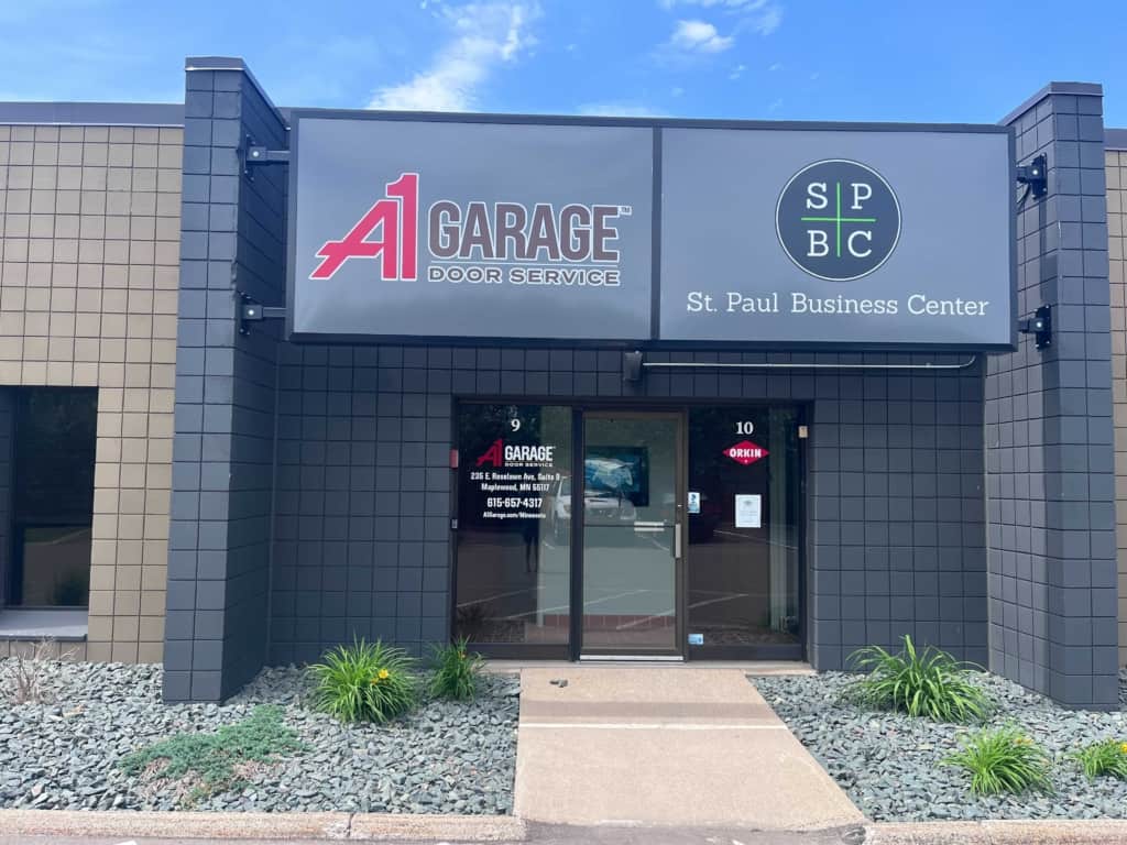 The storefront features signage for a garage door repair business called A1 Garage Door Service, alongside another sign for St. Paul Business Center, with a glass door entrance flanked by small landscaped plants and a clear blue sky overhead.